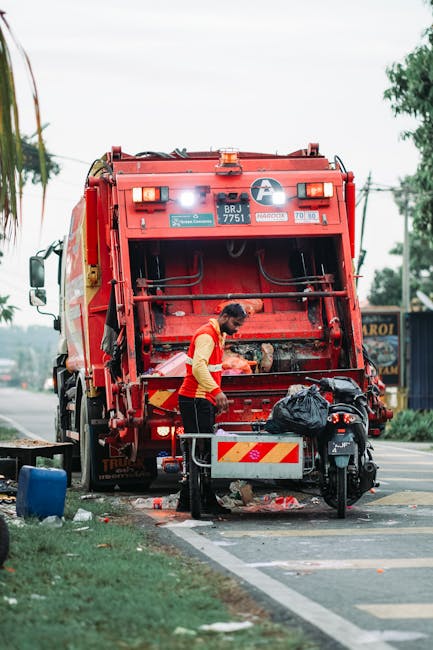 Two municipal workers wearing blue uniforms, yellow and black safety helmets, and pink gloves are seen on a city street, clearing green waste and scattered debris from the sidewalk. One worker is push
