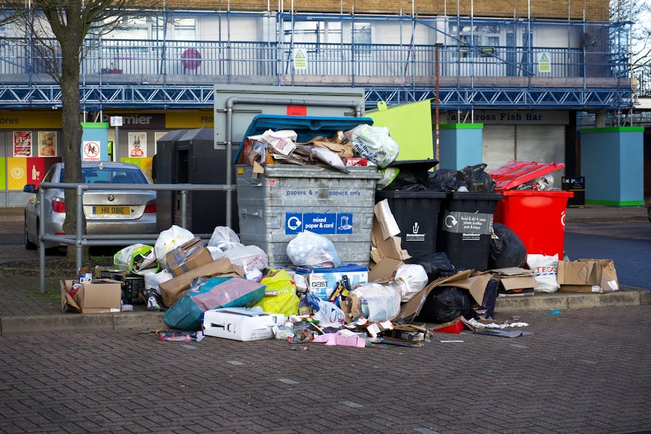 A pile of overflowing waste and rubbish bags situated next to a roadside curb in an urban area. The collection includes cardboard boxes, plastic bags, paper, and packaging materials scattered on the p