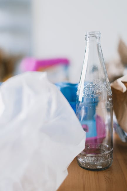 A clear glass soda bottle with embossed branding, positioned vertically on a wooden surface, partially surrounded by crumpled white paper and other miscellaneous waste materials. The bottle's surface